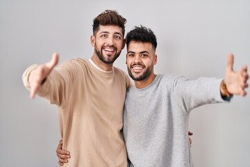Young homosexual couple standing over white background looking at the camera smiling with open arms for hug. cheerful expression embracing happiness. - obrazy, fototapety, plakaty