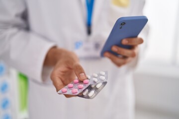 Young blond man pharmacist holding pills using smartphone at pharmacy