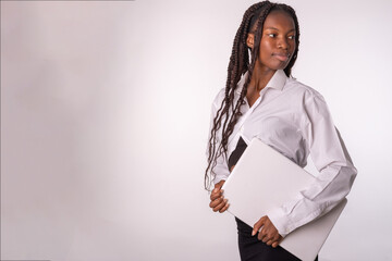 Cuerpo entero de una joven exitosa mujer de color de negocios empleada que usa una camisa blanca y una falda negral con una computadora portátil cerrada y una mirada a la derecha con el fondo blanco.
