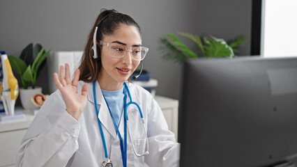 Young beautiful hispanic woman doctor doing video call with computer and headphones at clinic