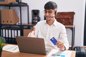 Young hispanic man working using computer laptop holding credit card smiling friendly offering handshake as greeting and welcoming. successful business.