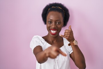 African woman with curly hair standing over pink background smiling doing talking on the telephone gesture and pointing to you. call me.