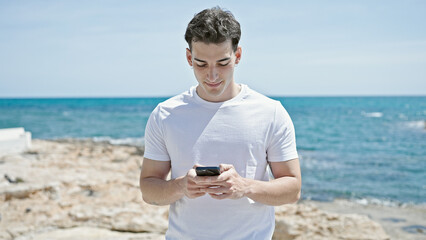 Young hispanic man using smartphone with serious expression at beach