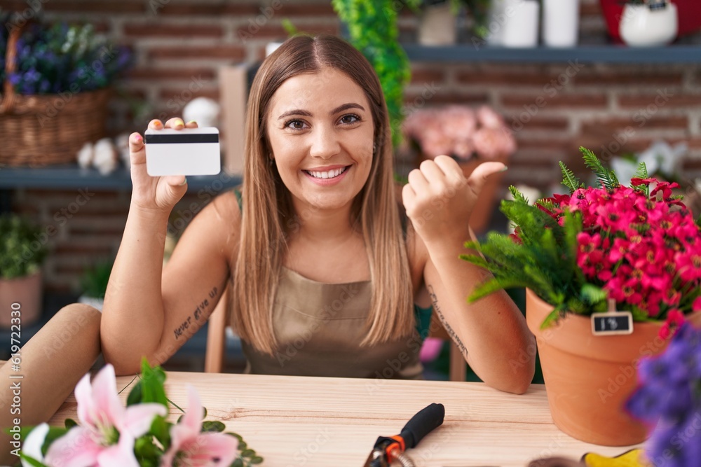 Poster young blonde woman working at florist shop holding credit card pointing thumb up to the side smiling