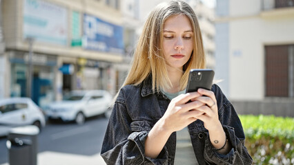 Young blonde woman using smartphone with serious face at street