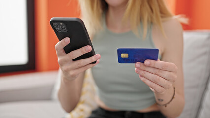 Young blonde woman shopping with smartphone and credit card sitting on sofa at home