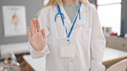 Young blonde woman doctor doing stop sign with hand at clinic