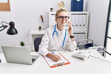 Young blonde woman doctor talking on telephone writing on document at clinic