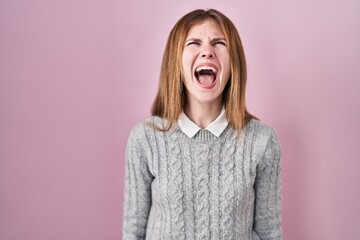 Beautiful woman standing over pink background angry and mad screaming frustrated and furious,...