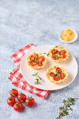 Unsweetened shortbread tartlets with feta cheese, cherry tomatoes and herbs on a white plate on a light concrete background. Unsweetened pastries,