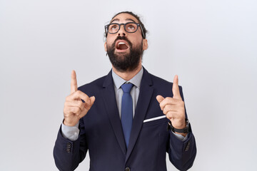 Hispanic man with beard wearing suit and tie amazed and surprised looking up and pointing with fingers and raised arms.