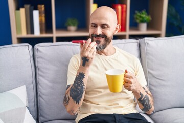 Young bald man talking on smartphone drinking coffee at home