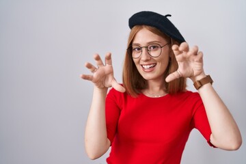 Young redhead woman standing wearing glasses and beret smiling funny doing claw gesture as cat, aggressive and sexy expression