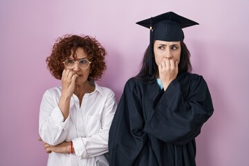 Hispanic mother and daughter wearing graduation cap and ceremony robe looking stressed and nervous...