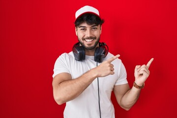 Hispanic man with beard wearing gamer hat and headphones smiling and looking at the camera pointing with two hands and fingers to the side. © Krakenimages.com
