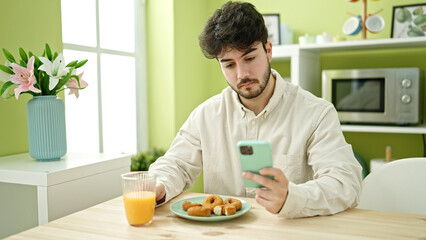 Young hispanic man having breakfast using smartphone at dinning room