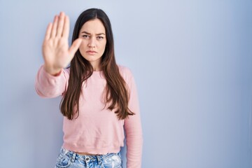 Fototapeta na wymiar Young brunette woman standing over blue background doing stop sing with palm of the hand. warning expression with negative and serious gesture on the face.