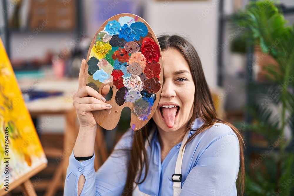 Wall mural young brunette woman covering face with painter palette sticking tongue out happy with funny express