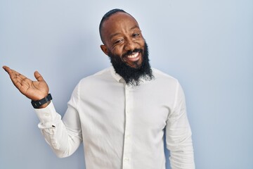 African american man standing over blue background smiling cheerful presenting and pointing with palm of hand looking at the camera.