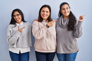 Mother and two daughters standing over blue background with a big smile on face, pointing with hand finger to the side looking at the camera.