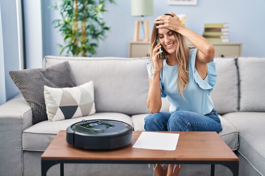 Young Hispanic Woman Sitting At Home By Vacuum Robot Speaking On The Phone Stressed And Frustrated With Hand On Head, Surprised And Angry Face