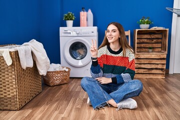 Young hispanic girl doing laundry smiling with happy face winking at the camera doing victory sign....