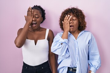 Two african women standing over pink background yawning tired covering half face, eye and mouth with hand. face hurts in pain.