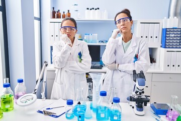 Fototapeta premium Young mother and daughter at scientist laboratory serious face thinking about question with hand on chin, thoughtful about confusing idea