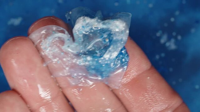 A soluble packet with blue and white powder, for a dishwasher, being held in my hand against a background of blue water where one has already dissolved. Close-up shot.