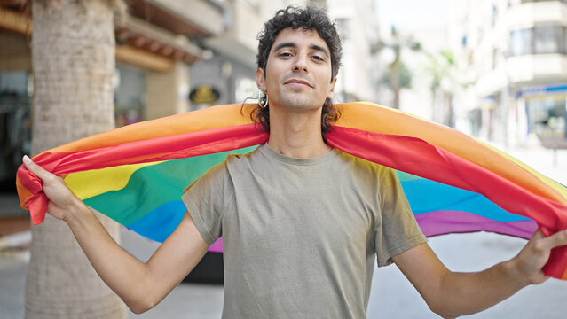 Young hispanic man holding rainbow flag with relaxed expression at street