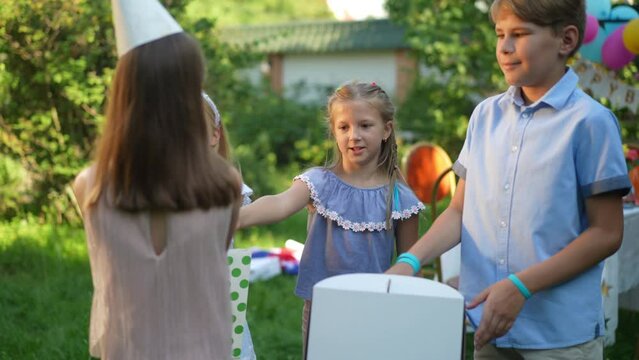 Positive kids walking to birthday girl passing presents smiling. Caucasian happy friends congratulating friend on holiday in sunny summer park