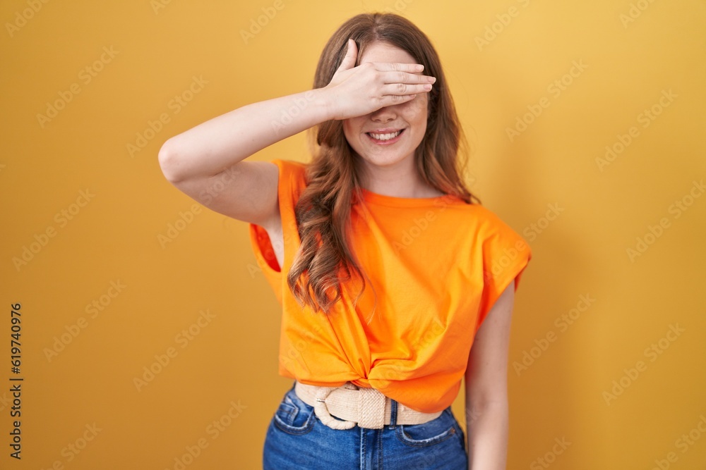 Poster caucasian woman standing over yellow background smiling and laughing with hand on face covering eyes