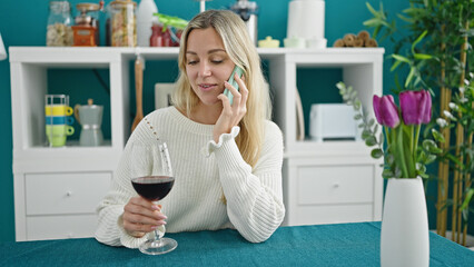 Young blonde woman drinking glass of wine talking on smartphone at dinning room