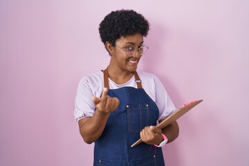 Young african american woman wearing professional waitress apron holding clipboard beckoning come...