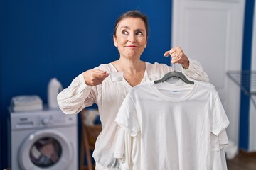 Middle age hispanic woman holding shirt on hanger and detergent powder smiling looking to the side...