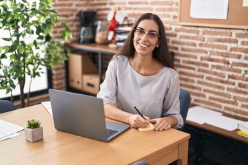 Young beautiful hispanic woman business worker using laptop writing on reminder paper at office