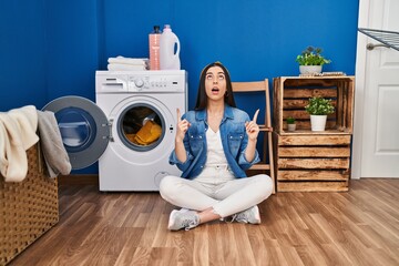 Hispanic woman doing laundry sitting on the floor amazed and surprised looking up and pointing with fingers and raised arms.