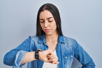 Hispanic woman standing over blue background checking the time on wrist watch, relaxed and confident