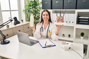 Hispanic doctor woman holding model of human anatomical skin and hair sticking tongue out happy with funny expression.