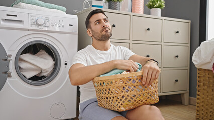 Young hispanic man sitting on floor tired holding wicker basket with clothes at laundry room