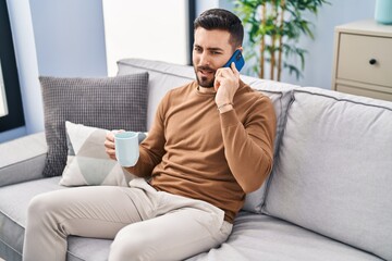 Fototapeta premium Young hispanic man drinking coffee and talking on the smartphone at home