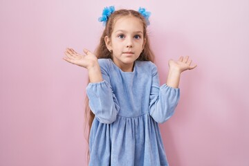 Young little girl standing over pink background clueless and confused expression with arms and hands raised. doubt concept.