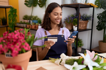 Young african american woman florist using smartphone holding credit card at flower shop