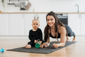 Fototapeta premium Joyful caucasian female holding plank pose on rubber mat near smiling little girl with sports bottle. Stylish mother building strong relationship with daughter while strengthening body at home.