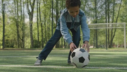 Cheerful African American boy walking on soccer field in park, putting ball on grass and preparing to kick it - Powered by Adobe