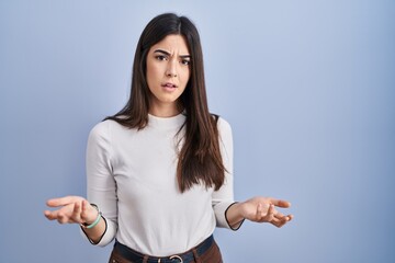 Young brunette woman standing over blue background clueless and confused with open arms, no idea...