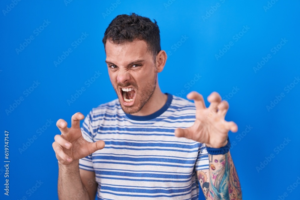 Poster Young hispanic man standing over blue background smiling funny doing claw gesture as cat, aggressive and sexy expression