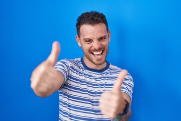 Young hispanic man standing over blue background approving doing positive gesture with hand, thumbs up smiling and happy for success. winner gesture.