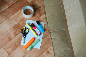 Open cardboard box with colorful markers, tape and scissors on the floor. Secondary consumption for playing with children