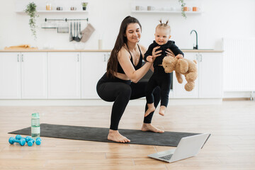 Smiling female holding cute girl with soft toy while working out squats to online video on kitchen background. Athletic mommy and child in yoga outfits enjoying benefits of quality time together.